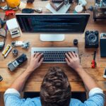 Mężczyzna kodujący na laptopie przy stole z starym sprzętem kamerowym wokół niego